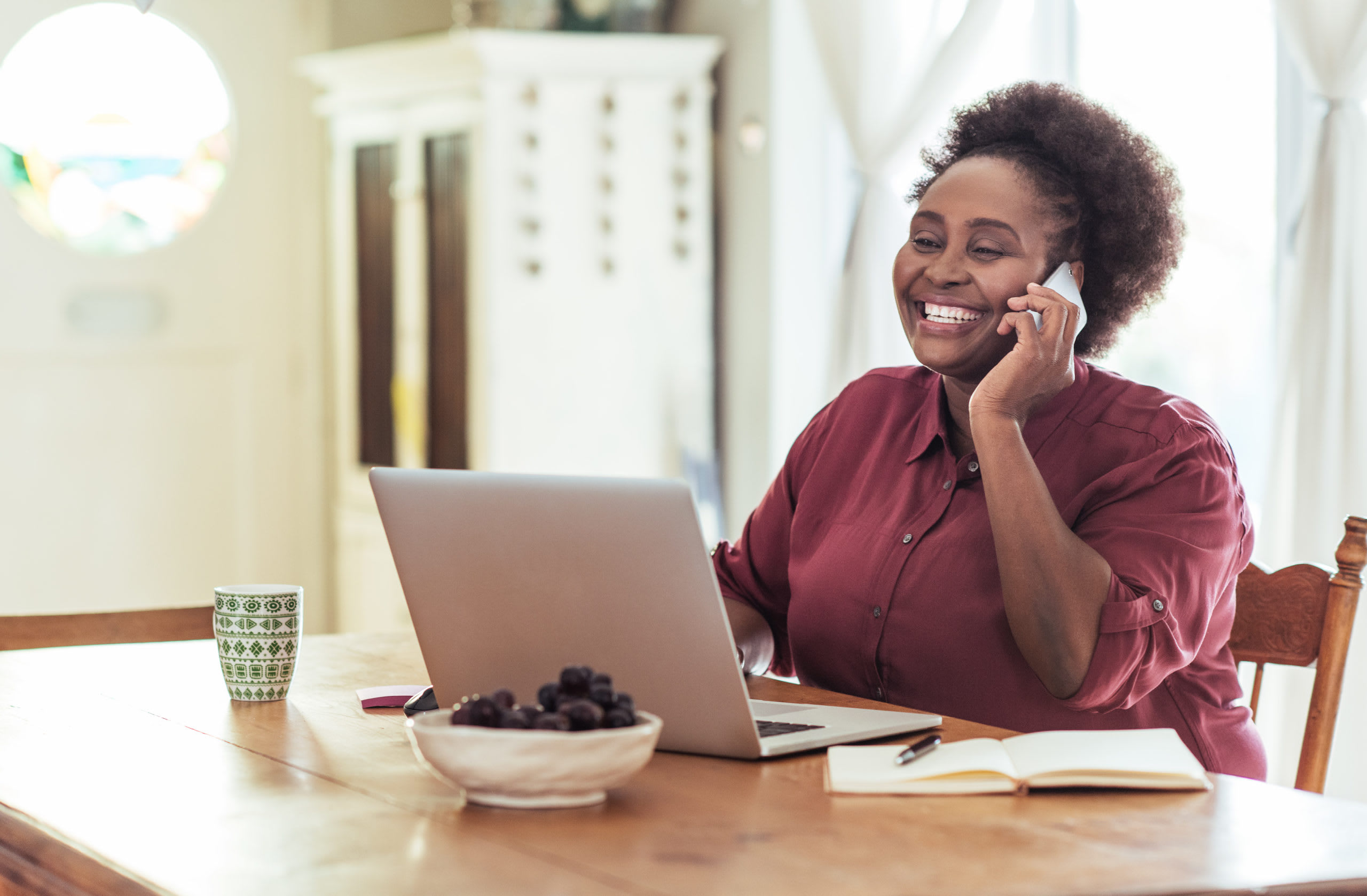 Person in red button-down talks on a cell phone and works on a laptop at a home desk.